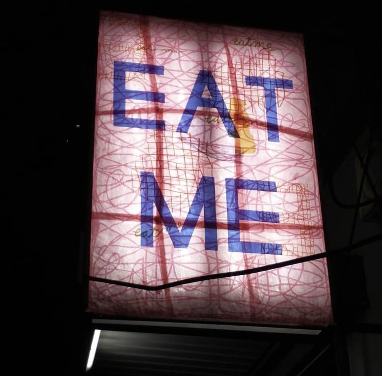 There is a photograph at night of a large backlit sign. In large blue letter on the sign is written “Eat Me” agains a transparent pink background. On top are darker pink straight and curved lines, as well as “eat me” written a few more times in cursive.