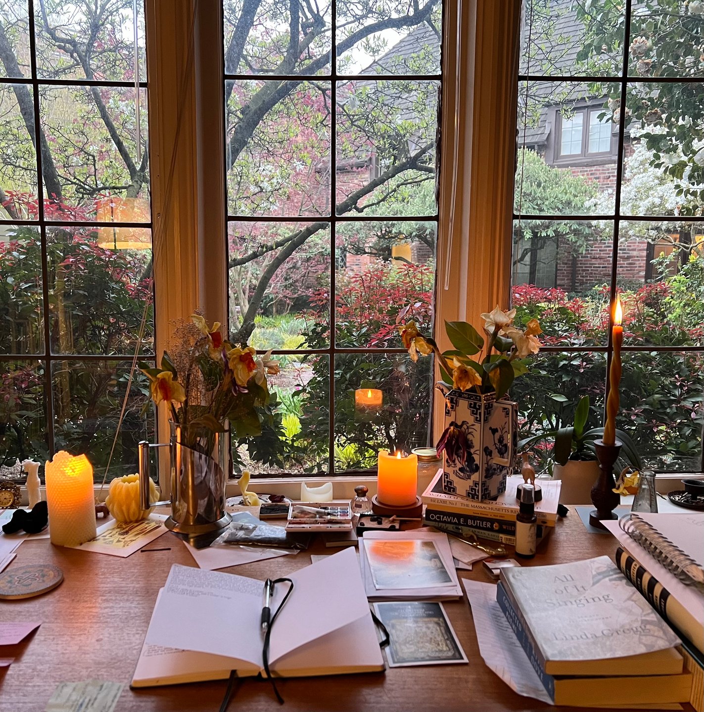 This is a photo of Bates' desk at dusk. The desk is covered in notebooks, postcards, and lit candles. A warm light comes from the candles and outside there are green trees and bushes.