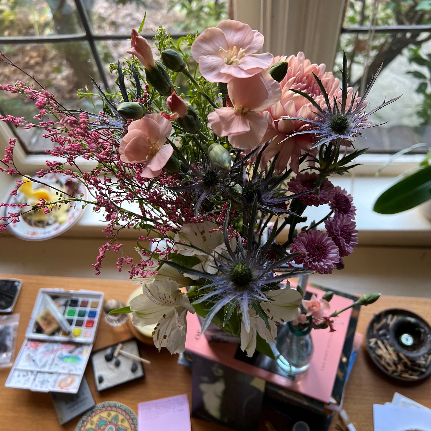 This photo is from above of a collection of pink wildflowers in a vase on the desk. In the background, there is incense and watercolors on the desk. 