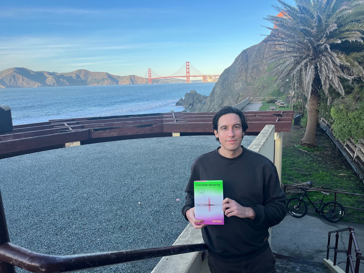 In this photograph, Josh is standing along the edge of a body of water, holding a copy of his novel. The Golden Gate Bridge is visible in the background.