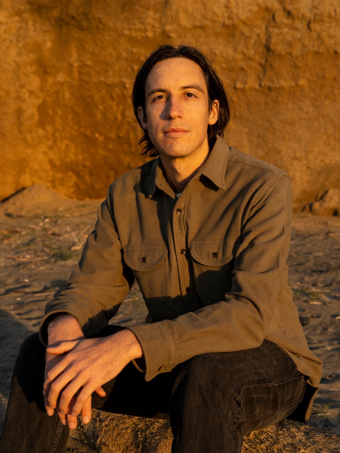 In this photograph, Josh is sitting on a beach. The light is yellow and warm, as if the photo was taken during golden hour.
