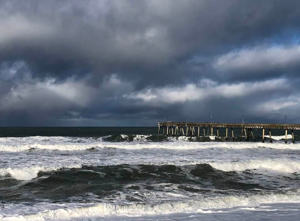 This picture shows a moody sky above a rough sea. Both the sky and sea are dark blue. On the right side of the photo, we see a pier stretching into the water