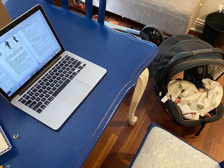 This photograph shows the author's laptop on a blue table with her baby sleeping in a baby carrier on the floor