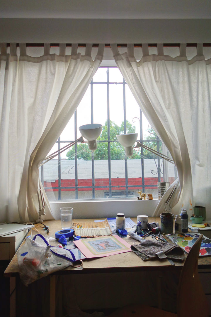 Photo of the artist's work desk, in front of a white-curtained window, surrounded by natural light and two lamps, with paints and supplies strewn throughout the desk space.
