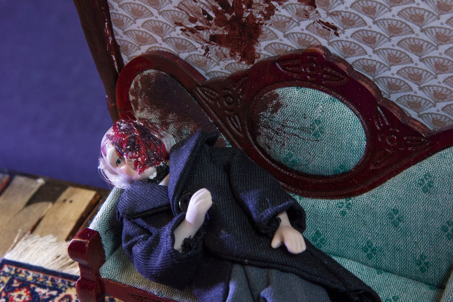 This photograph is a close up of the doll reclining on the couch. We see the gouge marks and blood spatters on his face and on the wall and the couch behind him.