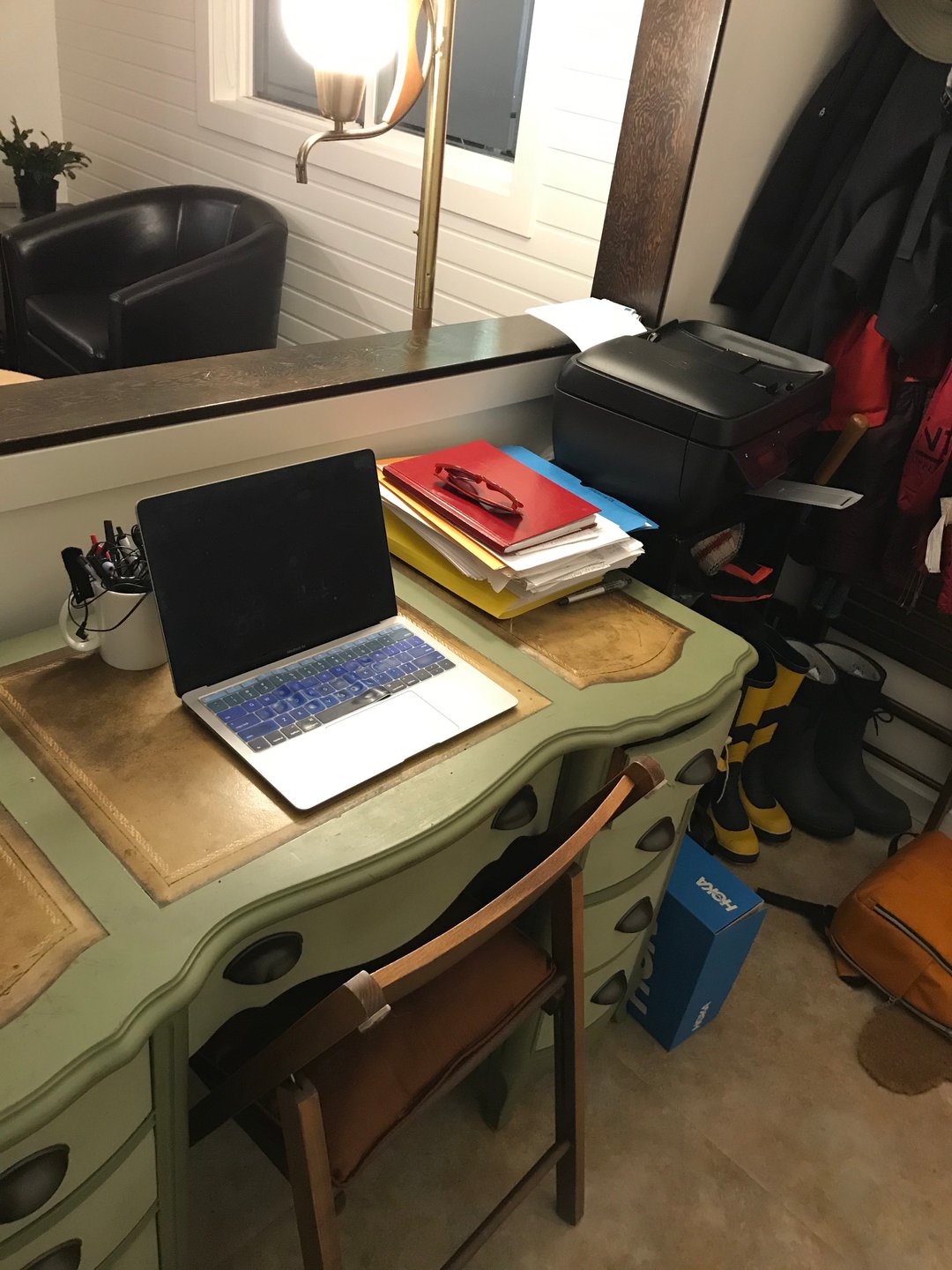 This photograph shows a green desk with a laptop on top of it. To the left, we see a printer with rainboots piled underneath it. A mirror behind the desk reflects a comfy chair and a lamp further back in the room.