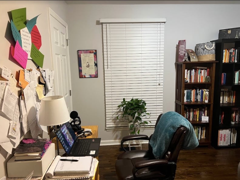 Destiny's office, with the pothos plant sitting in front of the window, bookshelves to the right of it. At the front of the photo (to the left hand side of the room), we see Destiny's desk with an office chair and a wall full of papers and notes