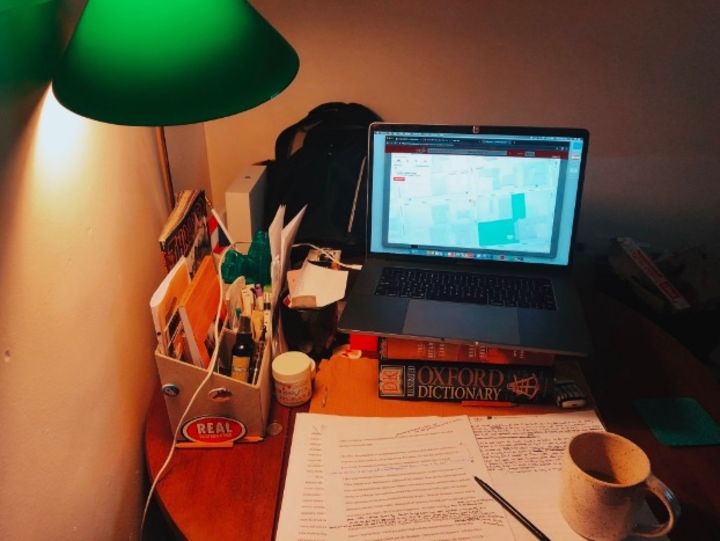 This photograph shows a laptop poised on top of books above a messy desk with a green lamp that casts a warm yellow glow