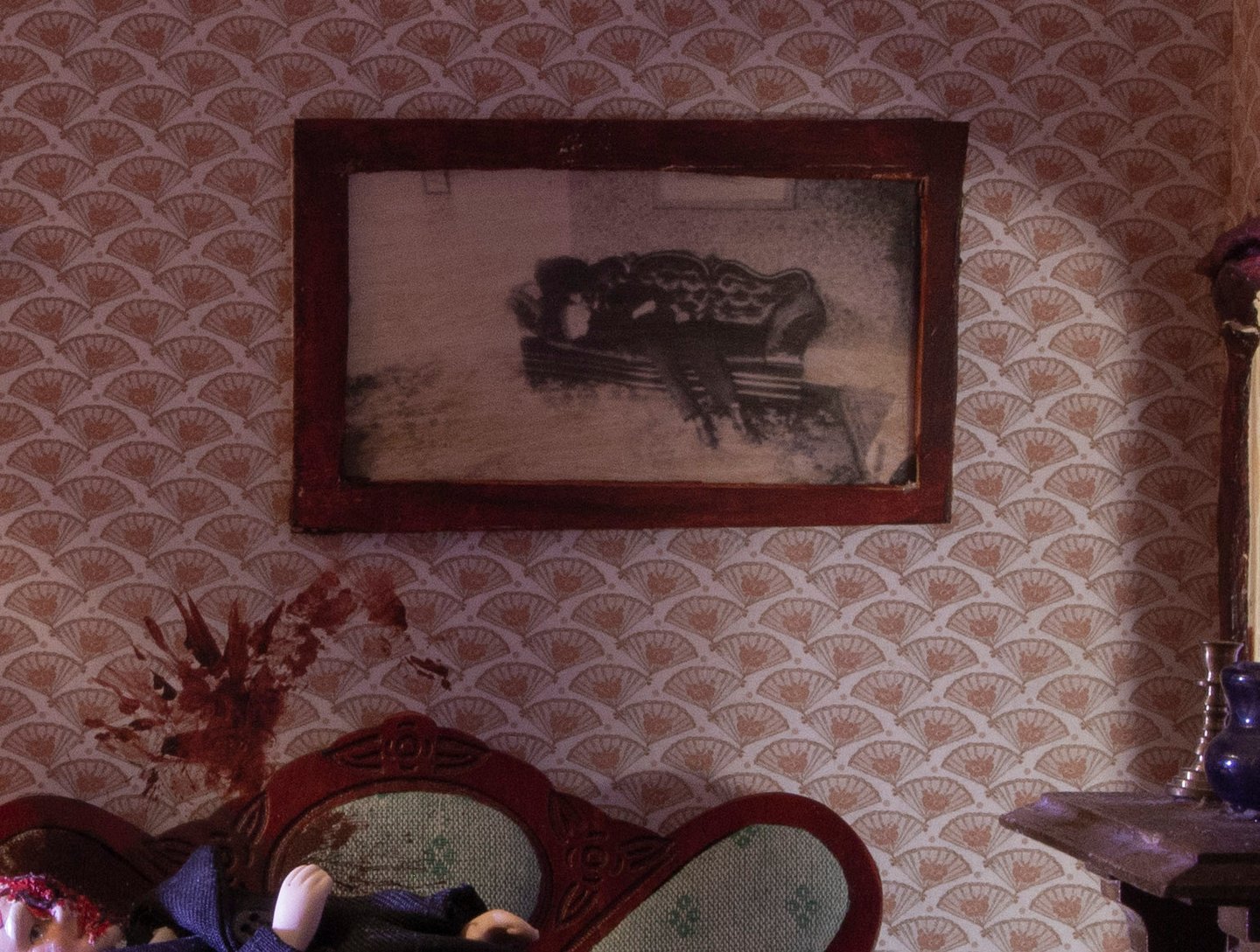 This photograph shows a close up of the wall of the dollhouse, where we see the picture that Zefyr described placing on the wall (of the father's own crime scene) above the doll laid out on the couch, blood spatters above his head.