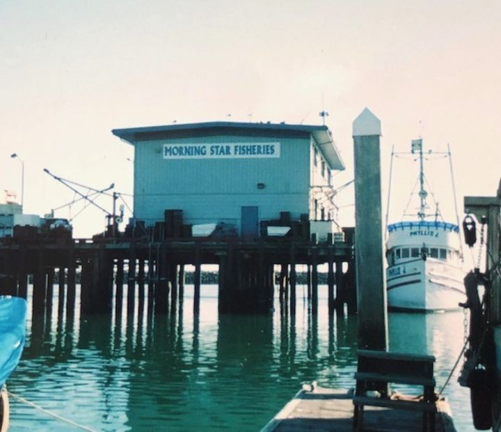 This photograph looks onto a pier. On top of the pier is a building with a sign that says "Morning Star Fisheries." We see a boat docked to the right of the building.